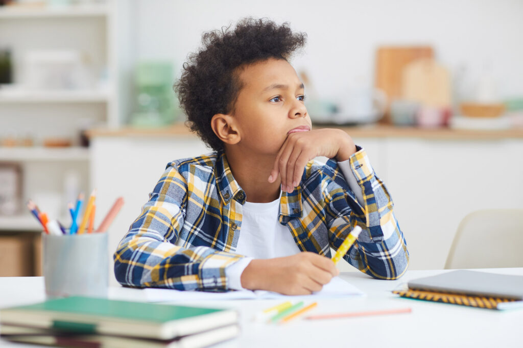 Ein kleiner Junge sitzt vor seinen Hausaufgaben und schaut verträumt in die Ferne
